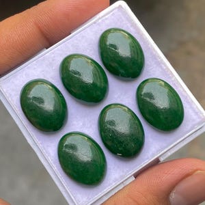 Puede incluir: Seis piedras preciosas verdes ovaladas y pulidas se muestran en una bandeja de plástico blanco. Las piedras preciosas tienen una superficie lisa y brillante y un color verde intenso y rico. Las piedras preciosas están dispuestas en dos filas de tres.