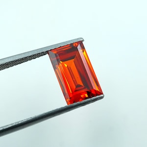 May include: A vibrant, rectangular-cut gemstone in a fiery orange hue is held by a pair of silver tweezers. The gem's facets reflect light, showcasing its clarity and brilliance. The background is a clean, bright white.