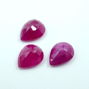 May include: Three faceted, pear-shaped gemstones in a vibrant, deep red hue. The gems are arranged on a white surface, showcasing their polished surfaces and intricate cuts. The gems are likely rubies.