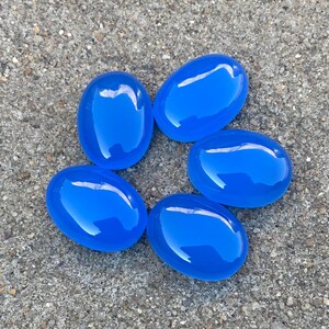 May include: Six smooth, oval-shaped, bright blue gemstones. The stones are arranged in a circle on a gray, textured surface.