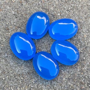 May include: Five smooth, oval-shaped blue gemstones arranged in a circle on a gray, textured surface.