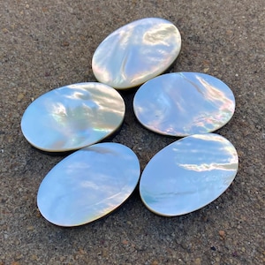 Könnte beinhalten: Fünf ovale Perlmutt-Cabochons mit irisierenden Farben. Die Cabochons sind in einem Kreis auf einer grauen, strukturierten Oberfläche angeordnet.