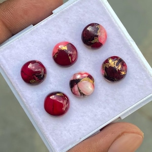 May include: Six round, polished gemstones in a white box. The stones are primarily red with gold and pink accents. The gemstones are held in a white box, and the hand of the person holding the box is visible.