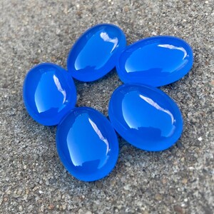 May include: Six smooth, oval-shaped blue gemstones. The stones are arranged in a circle on a gray, textured surface.