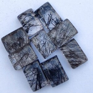 Peut inclure: Une collection de neuf cabochons en quartz clair avec des inclusions noires. Les cabochons sont rectangulaires et ont une surface lisse et polie.
