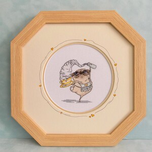 May include: Cross-stitch artwork of a pug wearing sunglasses, a hat, and a swimsuit, holding a drink. The artwork is framed in a light wood octagonal frame, with a cream-colored mat and a decorative heart border.