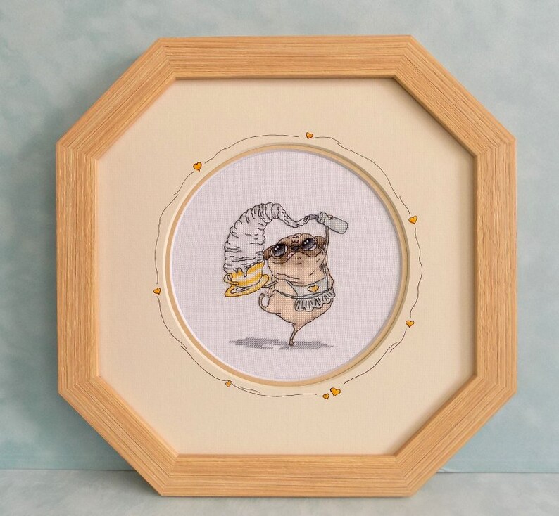 May include: Cross-stitch artwork of a pug wearing sunglasses, a hat, and a swimsuit, holding a drink. The artwork is framed in a light wood octagonal frame, with a cream-colored mat and a decorative heart border.