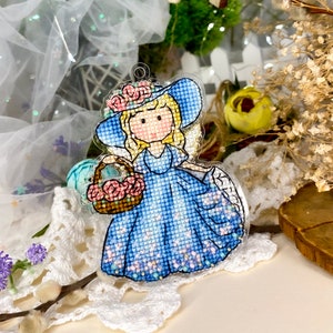 May include: A blue and white cross-stitch ornament of a girl wearing a blue hat with pink flowers and a blue dress holding a basket of pink flowers.