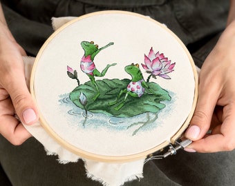 May include: Cross-stitch embroidery hoop with a design of two green frogs wearing pink and white swimsuits sitting on a green lily pad with a pink lotus flower.