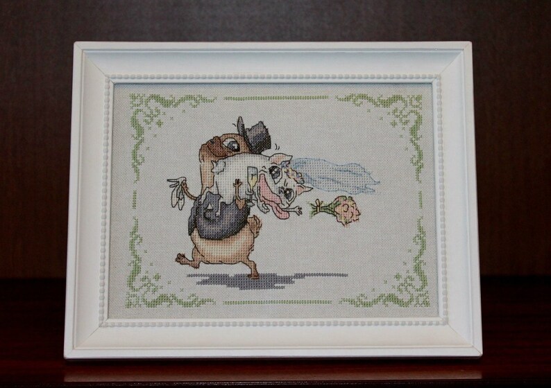 Peut inclure: Une broderie au point de croix encadr&eacute;e en blanc, repr&eacute;sentant une sc&egrave;ne de mariage de dessin anim&eacute; avec un chien brun portant un smoking portant un chat blanc portant une robe blanche et un voile.