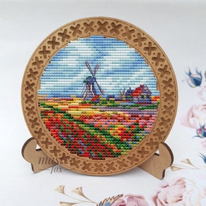 May include: A wooden round frame with a cross-stitch design of a windmill and a field of colorful tulips. The frame is on a wooden stand.