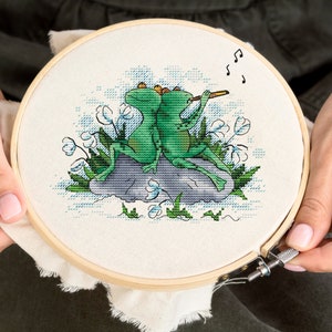 Könnte beinhalten: Ein Kreuzstich-Stickrahmen mit einem Motiv von zwei grünen Fröschen, die auf einem Stein sitzen, einer spielt Flöte. Die Frösche sind von weißen Blumen und blauem und grünem Laub umgeben. Das Motiv ist auf weißem Hintergrund.
