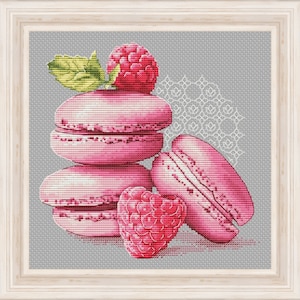 Op de afbeelding: Een stapel roze macarons met een framboos erop, omringd door een wit kantpatroon op een grijze achtergrond.