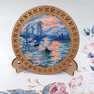 May include: A circular wooden frame with a cross-stitch design of a sunset over a lake. The frame is decorated with a repeating pattern of crosses and is mounted on a small wooden stand.