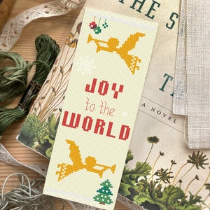 May include: A cream-coloured bookmark with a cross-stitch design featuring a yellow angel with a trumpet, a green Christmas tree, and the text "JOY to the WORLD" in red.