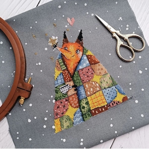 May include: A cross-stitch pattern of a fox wearing a colourful patchwork quilt. The fox is holding a cup and has a happy expression. The background is a grey fabric with white dots.