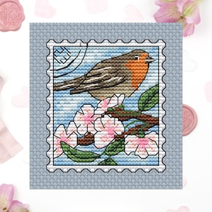 May include: A cross-stitch pattern of a robin perched on a branch with pink blossoms. The robin is brown, orange, and gray. The background is a blue sky with white clouds. The pattern is framed by a white border with a red stamp.