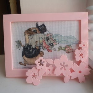 Peut inclure: Une broderie au point de croix encadr&eacute;e de rose repr&eacute;sentant deux animaux de dessin anim&eacute; se mariant. Le mari&eacute; est un animal brun portant un chapeau haut de forme et la mari&eacute;e est un animal blanc portant un voile. Le cadre est d&eacute;cor&eacute; de fleurs en plastique roses.
