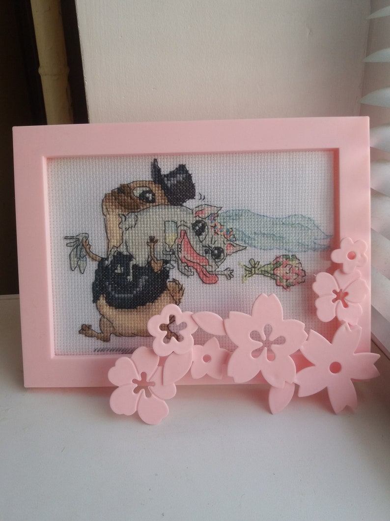 Peut inclure: Une broderie au point de croix encadr&eacute;e de rose repr&eacute;sentant deux animaux de dessin anim&eacute; se mariant. Le mari&eacute; est un animal brun portant un chapeau haut de forme et la mari&eacute;e est un animal blanc portant un voile. Le cadre est d&eacute;cor&eacute; de fleurs en plastique roses.
