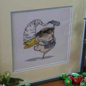 May include: Framed cross-stitch art featuring a pug wearing sunglasses and an apron, balancing a large ice cream cone. The artwork is in a light-colored frame with a blue border. The pug is beige, with black accents.