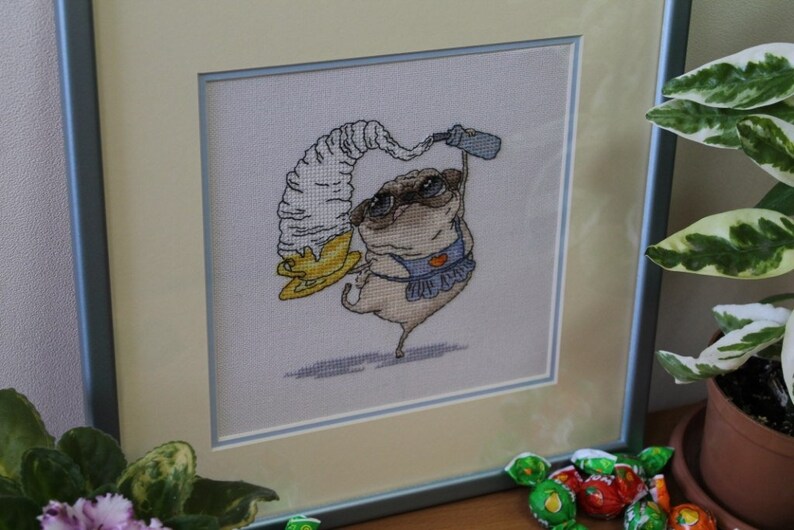 May include: Framed cross-stitch art featuring a pug wearing sunglasses and an apron, balancing a large ice cream cone. The artwork is in a light-colored frame with a blue border. The pug is beige, with black accents.