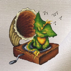 May include: Cross-stitch design of a green dragon listening to music on a vintage gramophone. The dragon is sitting on the gramophone, with musical notes floating above. The gramophone is brown and gold, and the background is white.