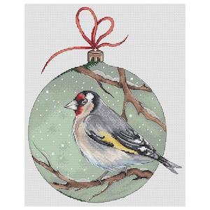 Puede incluir: Un adorno navideño verde con una cinta roja y un pequeño pájaro posado en una rama dentro. El adorno tiene un fondo nevado y un pájaro blanco, amarillo y gris con la cabeza roja.