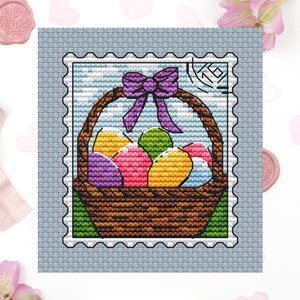 May include: A cross-stitch pattern of a brown wicker basket filled with colorful Easter eggs. A purple bow sits on top of the basket. The basket is framed by a blue and white postage stamp.