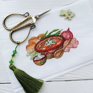 May include: A cross-stitch embroidery of a bowl of borscht, a traditional Ukrainian soup, with sour cream, onions, and bread. The embroidery is on white Aida cloth and is surrounded by green beads and a green tassel.