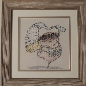 May include: Cross-stitch artwork of a pug wearing sunglasses and a frilly apron, balancing a large swirl of whipped cream and a bottle. The artwork is framed in a light-colored wooden frame with a white mat.