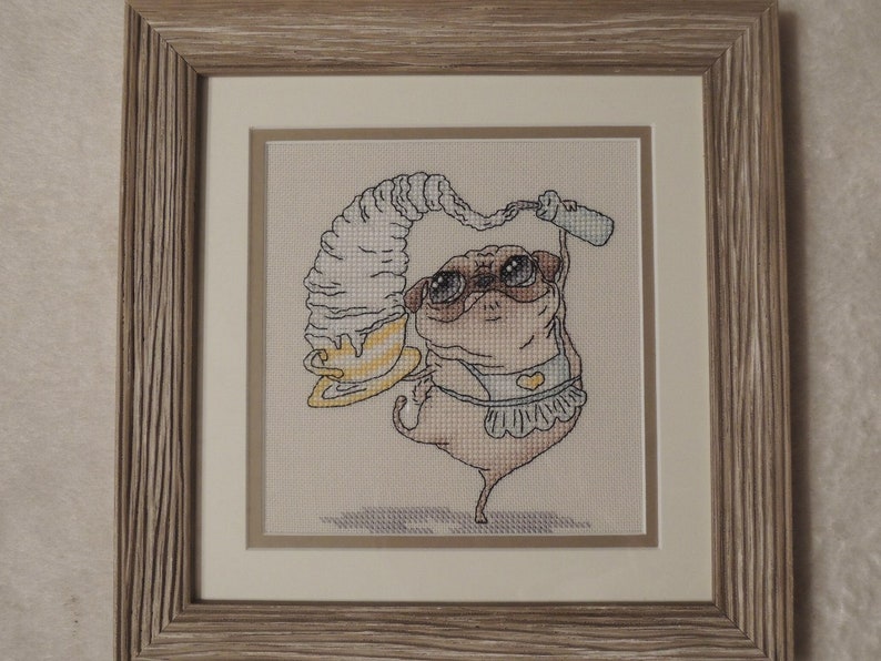 May include: Cross-stitch artwork of a pug wearing sunglasses and a frilly apron, balancing a large swirl of whipped cream and a bottle. The artwork is framed in a light-colored wooden frame with a white mat.