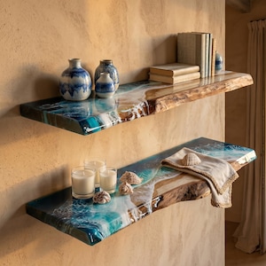Op de afbeelding: Twee houten planken met levendige randen en een harsachtig oceaanontwerp. De bovenste plank bevat boeken en blauw-witte vazen. De onderste plank heeft kaarsen, schelpen en een gevouwen linnen doek. De planken zijn aan een beige muur bevestigd.