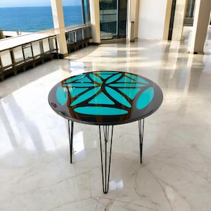 Puede incluir: Una mesa redonda con una tapa negra y una incrustaci&oacute;n de resina epoxi azul turquesa. La mesa tiene un dise&ntilde;o geom&eacute;trico y est&aacute; sostenida por cuatro patas de horquilla negras.