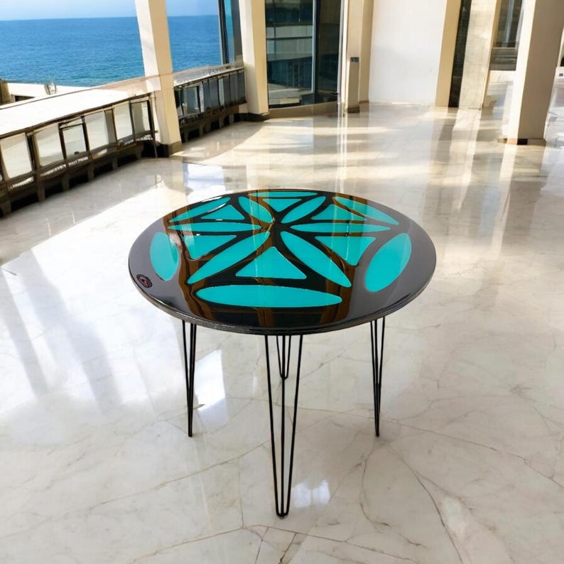 Puede incluir: Una mesa redonda con una tapa negra y una incrustaci&oacute;n de resina epoxi azul turquesa. La mesa tiene un dise&ntilde;o geom&eacute;trico y est&aacute; sostenida por cuatro patas de horquilla negras.