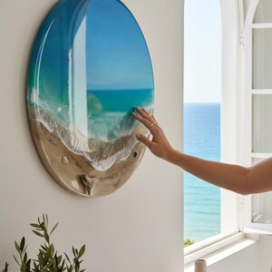 K&ouml;nnte beinhalten: Ein rundes Wandkunstwerk, das eine Strandszene mit blauem und t&uuml;rkisfarbenem Wasser, wei&szlig;en Wellen und einem sandigen Ufer zeigt. Das Kunstwerk ist an einer wei&szlig;en Wand befestigt. Eine Hand ber&uuml;hrt das Kunstwerk. Eine kleine Pflanze und dekorative Vasen befinden sich im Vordergrund.