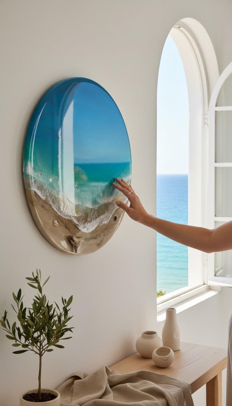 K&ouml;nnte beinhalten: Ein rundes Wandkunstwerk, das eine Strandszene mit blauem und t&uuml;rkisfarbenem Wasser, wei&szlig;en Wellen und einem sandigen Ufer zeigt. Das Kunstwerk ist an einer wei&szlig;en Wand befestigt. Eine Hand ber&uuml;hrt das Kunstwerk. Eine kleine Pflanze und dekorative Vasen befinden sich im Vordergrund.