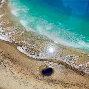 K&ouml;nnte beinhalten: Ein rundes Holzkunstwerk, das eine Strandszene mit einem blauen und t&uuml;rkisfarbenen Ozean, wei&szlig;en, sch&auml;umenden Wellen und einem sandigen Ufer darstellt. Ein kleiner, blauer und klarer Glasstein liegt auf dem Sand. Das Kunstwerk hat ein gl&auml;nzendes Finish.