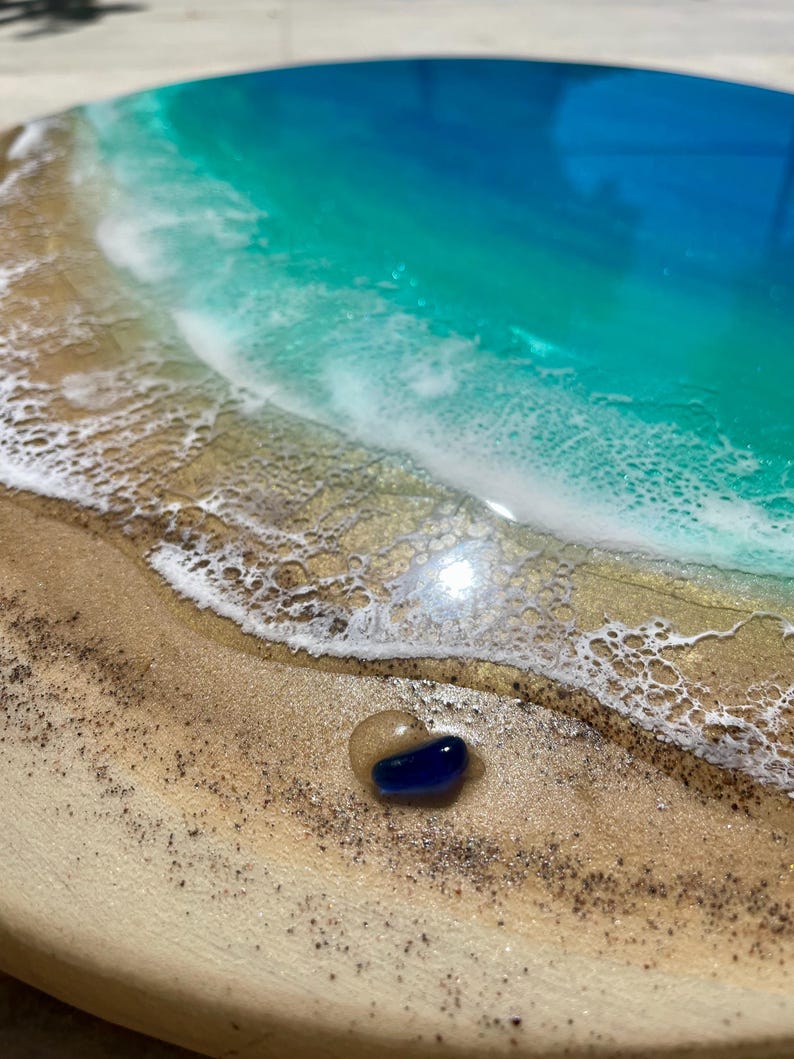 K&ouml;nnte beinhalten: Ein rundes Holzkunstwerk, das eine Strandszene mit einem blauen und t&uuml;rkisfarbenen Ozean, wei&szlig;en, sch&auml;umenden Wellen und einem sandigen Ufer darstellt. Ein kleiner, blauer und klarer Glasstein liegt auf dem Sand. Das Kunstwerk hat ein gl&auml;nzendes Finish.