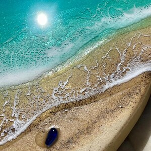 K&ouml;nnte beinhalten: Harzkunstwerk, das eine Strandszene in T&uuml;rkis- und Goldt&ouml;nen zeigt. Ein blauer Glasstein liegt am sandigen Ufer, mit schaumigen wei&szlig;en Wellen und einer hellen Sonne. Das St&uuml;ck hat ein K&uuml;stenmotiv, das an einen sonnigen Strand erinnert.