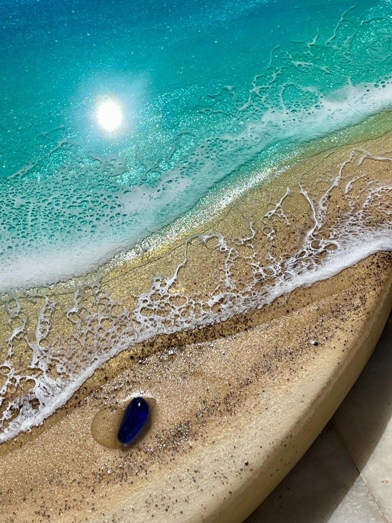 K&ouml;nnte beinhalten: Harzkunstwerk, das eine Strandszene in T&uuml;rkis- und Goldt&ouml;nen zeigt. Ein blauer Glasstein liegt am sandigen Ufer, mit schaumigen wei&szlig;en Wellen und einer hellen Sonne. Das St&uuml;ck hat ein K&uuml;stenmotiv, das an einen sonnigen Strand erinnert.