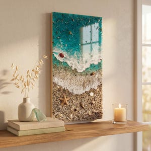 May include: A rectangular resin art piece depicting a beach scene. The top section shows turquoise water with white foamy waves. The bottom section features sand, seashells, and a starfish. The artwork is framed in a light-colored wood.