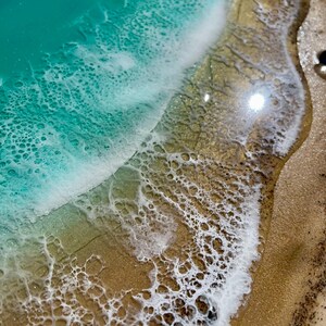 K&ouml;nnte beinhalten: Nahaufnahme eines Harzkunstwerks mit Meeresmotiv. Das Kunstwerk zeigt einen t&uuml;rkisblauen Ozean mit wei&szlig;en, schaumigen Wellen, die auf einen Sandstrand treffen. Der Strand hat einen goldenen Farbton mit strukturierten Details.