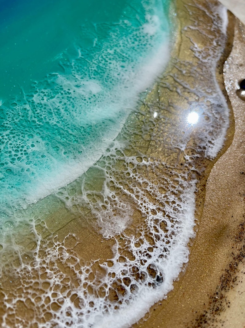 K&ouml;nnte beinhalten: Nahaufnahme eines Harzkunstwerks mit Meeresmotiv. Das Kunstwerk zeigt einen t&uuml;rkisblauen Ozean mit wei&szlig;en, schaumigen Wellen, die auf einen Sandstrand treffen. Der Strand hat einen goldenen Farbton mit strukturierten Details.