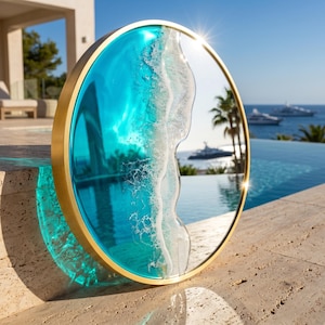 Könnte beinhalten: Ein runder Wandspiegel mit einem goldfarbenen Metallrahmen und einer dekorativen Harzeinlage, die eine türkisfarbene Meereswelle mit weißem Gischt-Effekt nachahmt. Die reflektierende Oberfläche spiegelt die umliegende Außenlandschaft wider.