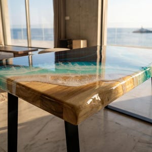 Op de afbeelding: Een rechthoekige tafel met een natuurlijke houten rand en een glanzend hars oppervlak. De hars toont een kustscène met zandbeige tinten, turkooisblauw water en witte schuimgolven die overgaan in een diep marineblauw oceaangedeelte.