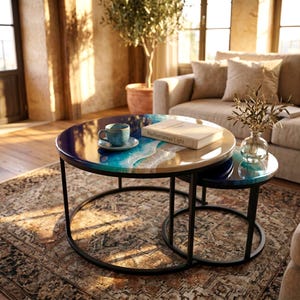 Peut inclure: Un ensemble de deux tables basses gigognes rondes avec des cadres en métal noir et des plateaux en résine arborant un motif de vagues océaniques bleu et blanc. Une tasse bleue sur soucoupe et un livre intitulé Mediterranean Light sont posés.