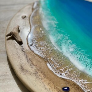 K&ouml;nnte beinhalten: Rundes Holzkunstwerk, das eine Strandszene darstellt. Das Kunstwerk zeigt einen sandigen Strand, sch&auml;umende wei&szlig;e Wellen und einen Farbverlauf aus T&uuml;rkis und blauem Wasser. Ein St&uuml;ck Treibholz und ein kleiner blauer Stein liegen am Strand.