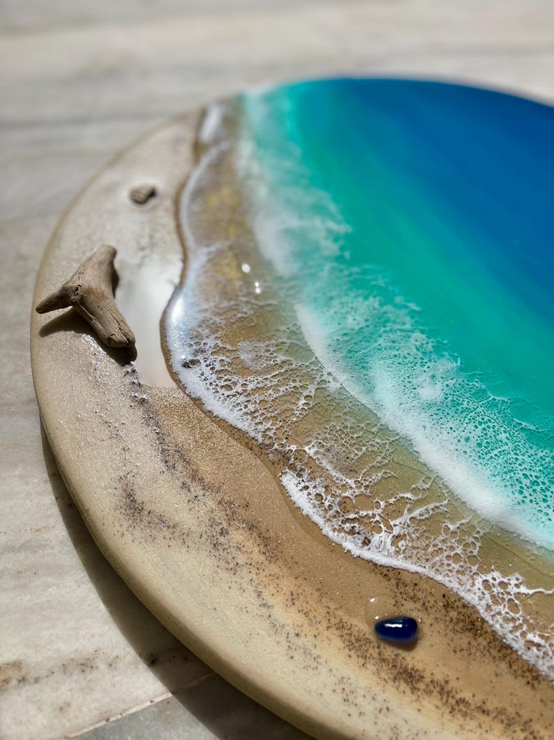 K&ouml;nnte beinhalten: Rundes Holzkunstwerk, das eine Strandszene darstellt. Das Kunstwerk zeigt einen sandigen Strand, sch&auml;umende wei&szlig;e Wellen und einen Farbverlauf aus T&uuml;rkis und blauem Wasser. Ein St&uuml;ck Treibholz und ein kleiner blauer Stein liegen am Strand.