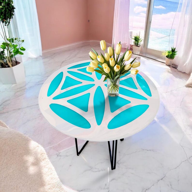 Puede incluir: Una mesa redonda blanca con un dise&ntilde;o geom&eacute;trico en azul turquesa. La mesa tiene una base de metal negro y un jarr&oacute;n de flores amarillas en la parte superior.