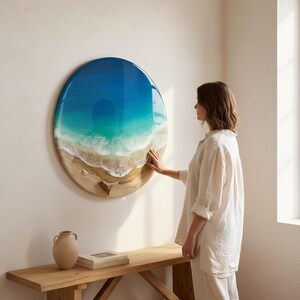 K&ouml;nnte beinhalten: Runde Wandkunst mit einer Strandlandschaft mit blauem und t&uuml;rkisfarbenem Wasser, wei&szlig;en Wellen und einem sandigen Ufer. Das Kunstwerk h&auml;ngt an einer wei&szlig;en Wand. Eine Person ber&uuml;hrt das Kunstwerk.